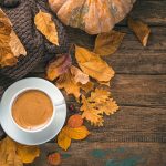 Photo shows an autumn-themed composition shot from above. On an old wooden table we see a cup of hot black coffee on a white saucer, surrounded by golden autumn leaves, a pumpkin and an comfy woollen sweater.