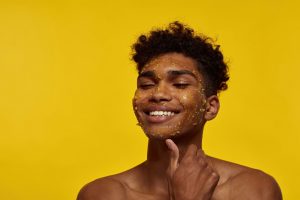 Head and shoulders photo of a young black man in front of a yellow background, applying a honey scrub to his face. He is smiling as he runs his thumb along his jaw.