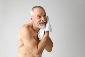 A mature caucasian man with a whte beard uses a flannel to clean his face.