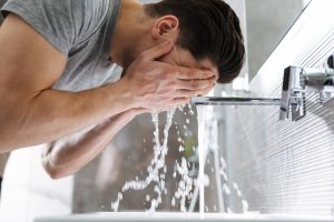Head and shoulders shot of a young caucasian man in a grey t-shirt in his bathroom. He is washing his face at the sink, his head down and face covered by his hands as he splashes his face with water.