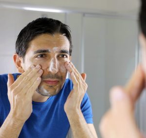 A man is shown reflected in his bathroom mirror, wearing a bright blue t-shirt and washing his face with a facial cleanser.