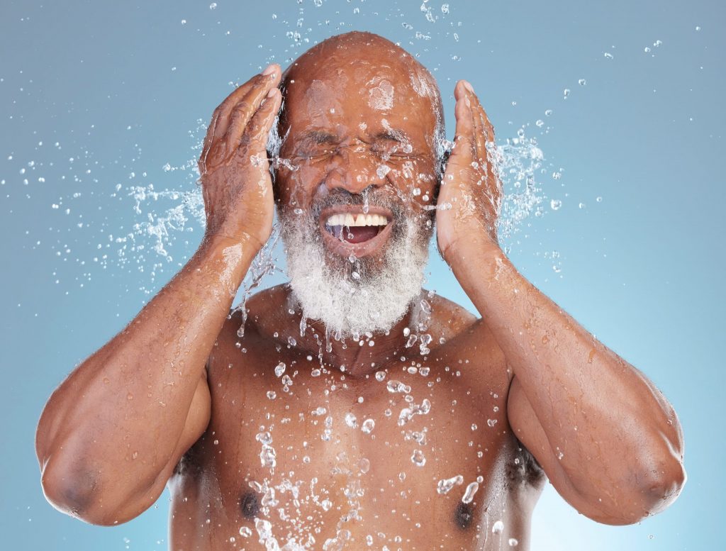 An older black man with a white beard is shown from the chest up, washing his face. His eyes are closed and he laughs as the water hits his skin in an exaggerated splash.