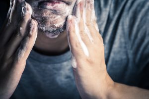 Extreme close up of a clean shaven caucasian man in a grey t-shirt massaging facial cleanser into his chin and cheeks. We only see his jaw and hands, and the cleanser is foaming as he rubs it in.