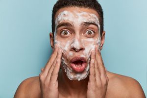 Portait of a young man looking surprised as he washes his face. His skin is covered in excessive foam from the facial cleanser and he clutches his face and glances open-mouthed at the camera in shock.