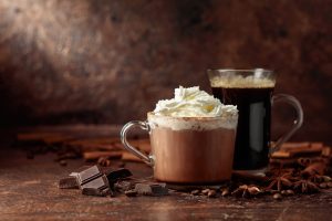 Photo shows a glass of hot chocolate with whipped cream and marshmallows, alongside a taller glass of strong black coffee. They are placed on a wooden surface with chunks of broken chocolate and cinnamon sticks around them, against a dark brown background.