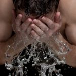 An athletic young man washing his face. He is facing us with his head down and splashing his face with water.