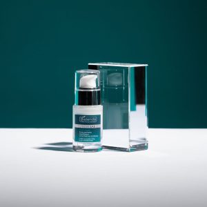 Anti-wrinkle eye cream from Bielenda, part of their men's skincare product line. Photo shows a 15ml glass bottle of eye cream reflected in a block of glass, against a bluey green background and with strong areas of light and shadow.