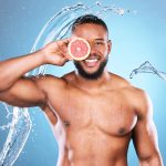 Portrait of a young black man, standing shirtless against a blue background. He is smiling broadly and holding a sliced grapefruit in front of his face, as he is splashed with water from both sides.