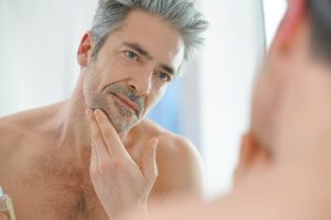 A head and shoulders portrait of an older man with silver hair, looking at himself in the mirror while he strokes his chin.