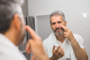 A smiling middle-aged man with greying hair and beard is in his bathroom. Dressed in a white t-shirt with a towel around his neck, he is shown applying moisturiser to his cheeks.