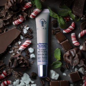 Product shot of Peppermint Mocha Lip Hydrator frm CIRCADIA. A small white tube of lip balm with a silver lid rests on a grey slate, surrounded by pieces of chocolate, green mint leaves, coffee beans, and red and white striped candy canes.