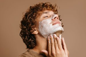Handsome young man with curly auburn hair in a brown t-shirt, shown from the side applying a white face mask to his cheeks and jaw. His head is tilted towards the top right of the picture and he is smiling slightly as he runs his hands over his neck.