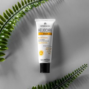 Product shot of Fluid Cream SPF from HELIOCARE. A white tube with yellow writing and a black cap is depicted against a grey background, circled by two green fronds of fern.
