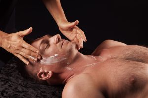 A handsome, middle-aged Causcasian man is receiving a face massage. He is depicted from the chest up, lying on a dark, fluffy towel. We can only see the therapist's hands - one hand is placed on the man's right temple, the other hand on the left side of his jaw. We can see from traces of product on the skin that cream has been applied to his neck, chin and cheeks. The image is dramtically lit against a black background.