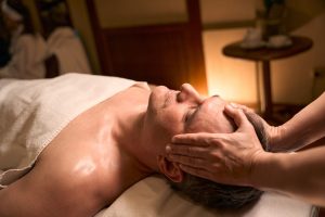 A middle-aged man is shown receiving a scalp massage in a spa. He is shown from the chest up, wrapped in a beige towel, lying on a treatment bed in a warmly lit room. We see the therapist's forearms and hands only, thumbs resting on the customer's forehead and fingers on his temples.