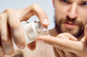 Close-up shot of a young, bearded man pumping moisturiser from a white bottle onto his finger. His hands and the bottle are in focus at the front of the picture, his face and chest are blurred against a light background.