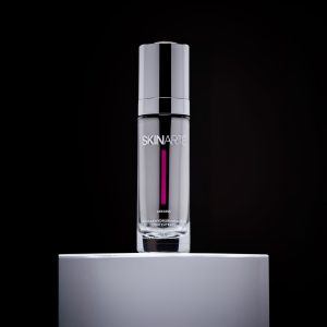 Product shot of Cellular Hydrorevitalizing Concentrate from SKINARTÉ. A luxurious-looking silver and glass bottle stands on top of a white pedestal, dramatically lit against a black background.