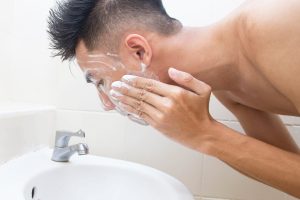 A young, shirtless Chinese man is leaning over a bathroom sink and washing his face with a foaming cleanser.