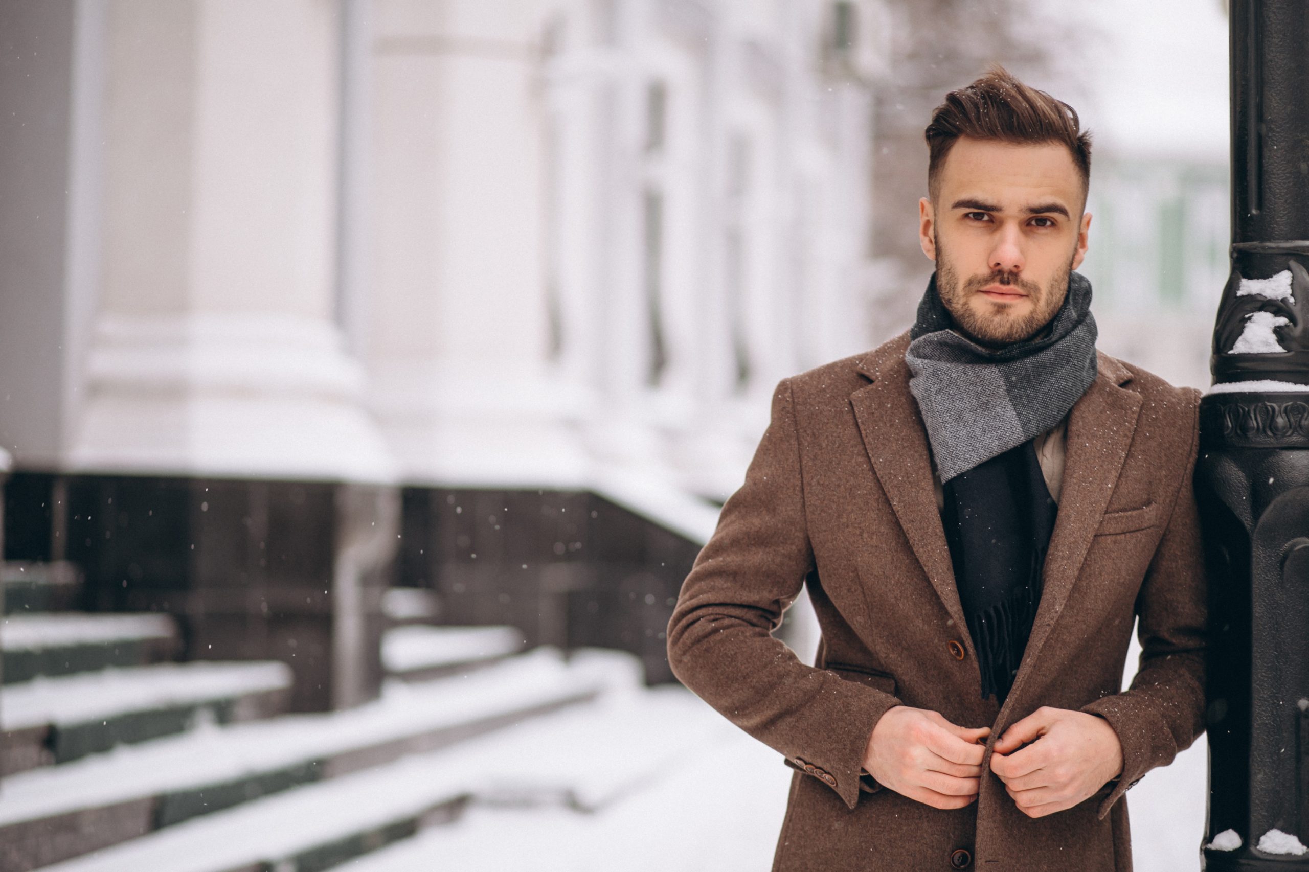 A young, bearded Caucasian man is leaning against a lampost in a snowy street. There is snow on the pavement and on the steps of the buildings behind him. He is shown from the waist up, smartly dressed in a brown coat and grey scarf. He is buttoning the coat against the elements, as snow falls around him.