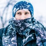 A head and shoulders shot of a young man in a frozen landscape. He is wrapped up in a winter coat, scarf and woolly hat, covered in snow, with just his nose and eyes peeking over the top of his scarf.